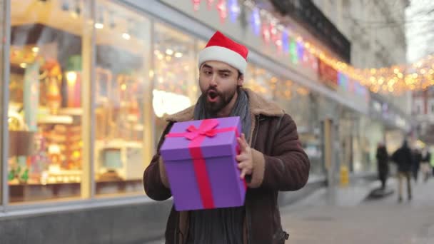 Joyful man in santa hat shaking with gift box on city street — Stockvideo