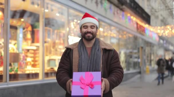 Smiling man in santa hat showing gift box on camera — Stockvideo