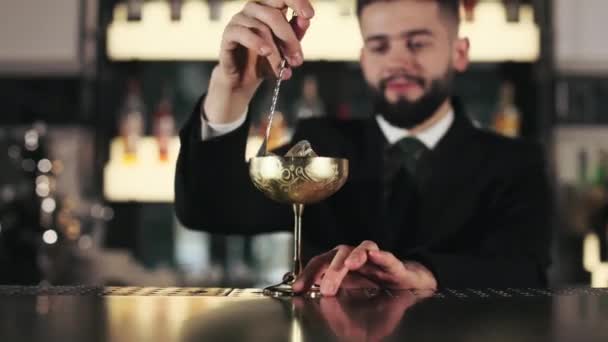 酒保精明能干 身穿时髦黑色西服 搅动酒精饮料 并在金黄色古董酒杯中加入冰块和长勺 历史鸡尾酒的配方 — 图库视频影像