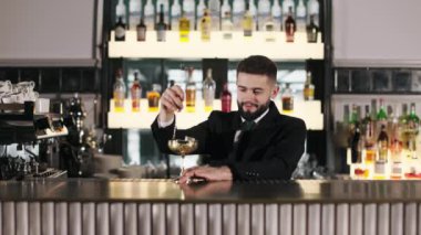 Profesyonel barmen siyah takım elbise ve kravat takıyor. Parlak altın şarap kadehinde buz küpleriyle kokteyl karıştırmak için uzun kaşık kullanıyor. Yakışıklı sakallı adam lüks restoranda alkollü içecek hazırlıyor..