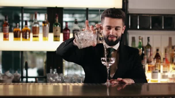 白种人英俊的酒保通过过滤将清凉的饮料倒入玻璃杯 一个留着胡子的年轻人 穿着黑色西服 打着领带 在奢侈品餐厅工作 — 图库视频影像