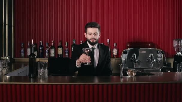 お酒のカクテルとバーカウンターのガラスに置くスタイリッシュな黒のスーツで白亜のバーマンの笑顔 豪華なレストランでドリンクを提供するハンサムな男の肖像 — ストック動画