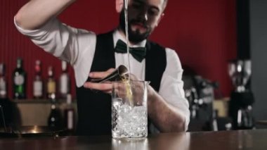 Deneyimli Kafkas barmen üniformalı alkollü barmen ölçü kabından buzlu cama içki dolduruyor. Yakışıklı sakallı adam tezgahta duruyor ve benzersiz bir tarif hazırlıyor..