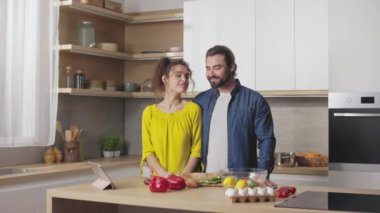 Gayri resmi giyinmiş güzel beyaz bir çiftin portresi. Mutfakta yan yana dururken gülümseyen ve kameraya bakan. Dijital tablet ve taze sebzeler..