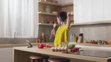 Mutfakta şarkı söyleyip dans ederken mikrofon olarak tahta kaşık kullanan kulaklıklı beyaz bir kadın. Evde eğlence ve zevkle yemek pişirmek..