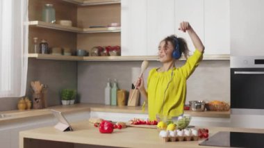 Joyful young woman with curly hair listening music in headphones while preparing fresh vegetable salad at home. Charming lady dancing and singing during cooking process on kitchen.