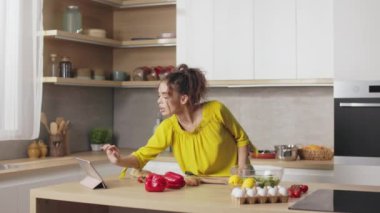 Parlak mutfakta yemek pişirirken dijital tablet kullanan gündelik kıyafetli çekici genç bir kadın. Kafkasyalı esmer taze sebzelerden sağlıklı salata hazırlıyor..