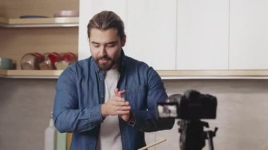 Sıradan giyinen yakışıklı sakallı adam parlak mutfakta yeni video bloğu çekiyor. Beyaz erkek yemek blogcusu yemek pişirirken kameraya konuşuyor ve el kol hareketi yapıyor..