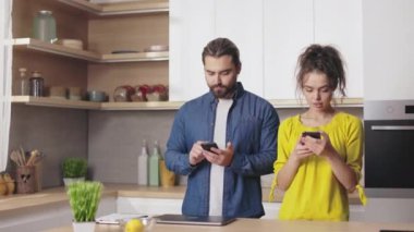 Evdeki modern akıllı telefonları kullanarak başlarını çeviren ve birbirlerine bakan güzel evli bir çift. Arka planda mutfak var. Teknoloji ve yaşam tarzı kavramı.