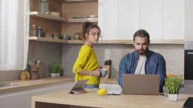 Handsome bearded man typing on wireless laptop while her charming wife standing near with cup of hot drink in hands. Lifestyles of caucasian family at home.