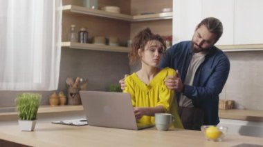 Caucasian man doing shoulder massage to his charming wife that working on laptop at kitchen table. Concept of freelance, relationship and support.