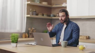 Yakışıklı sakallı adam evde çalışırken video konferansı için modern laptop kullanıyor. Kafkasyalı serbest çalışan mutfak masasında oturuyor, konuşuyor ve el kol hareketi yapıyor..