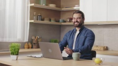 Joyful caucasian man in casual wear gesturing thumbs up while working on modern laptop at kitchen desk. Concept of freelance, success and technology.