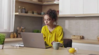 Focused caucasian woman with curly hair sitting at kitchen tablet and using wireless laptop. Beautiful brunette working remotely from cozy home.