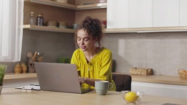 Pleasant caucasian woman with curly brown hair working on portable laptop among bright domestic kitchen. Female freelancer with modern computer at home.
