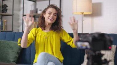 Günlük giysiler içinde kadın medya blogcusu evde sosyal ağ için canlı video yayınlarken kameraya el sallıyor. Heyecanlı kadın, profesyonel kamera kullanarak kanal takipçilerini selamlıyor.