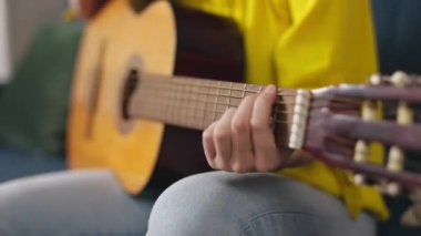 Close up of womans hands playing classic wooden guitar while sitting on couch at home. Musical instrument for recreation and hobby passion.