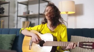 Young female player sitting on sofa in living room and performing song using favorite musical instrument. Beautiful woman with curly hair playing acoustic wooden guitar and singing alone at home.