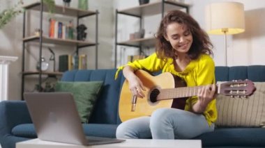 Beyaz tenli yetenekli bir kadın, evde pratik yaparken gitar çalıyor ve dizüstü bilgisayarda ders izliyor. Boş zaman harcamaları ve çevrimiçi eğitim kavramı.