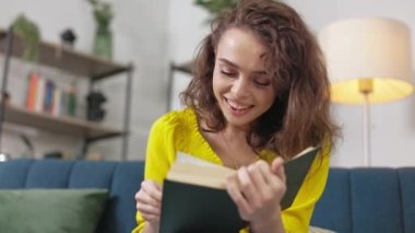 Çekici, kıvırcık saçlı, kanepede otururken ilginç kitaplar okuyan genç bir kadın. Yaratıcı edebiyat kavramı ve boş zaman geçirme.