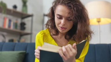 Rahat koltukta kitap elinde dinlenen gündelik giysili çekici genç bayan. Heyecanlı kıvırcık saçlı kadın evde boş zamanlarında büyüleyici edebiyat kitapları okuyor..