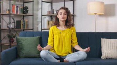 Gorgeous young woman in casual clothes keeping eyes closed and practicing peace of mind meditation at home. Caucasian female improving inner balance while sitting in lotus pose on couch.
