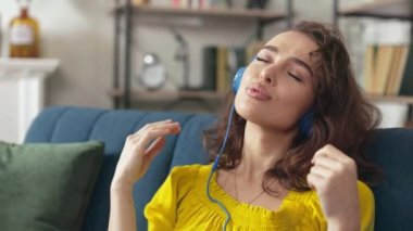 Charming woman in headphones keeping eyes closed and dancing with hands while listening favorite music at home. Caucasian female sitting on sofa in modern living room and enjoying calm melody.