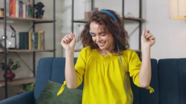 Female music lover in headphones resting alone on comfy sofa and listening favorite song. Pleasant woman dancing actively with hands and waving with her curly brown hair.