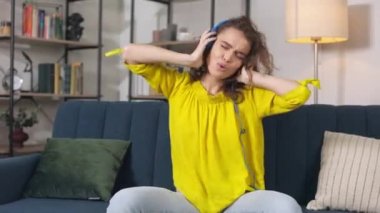 Cheerful young woman in headphones sitting on couch, listening music and moving actively. Happy caucasian female enjoying popular soundtrack. Domestic entertainments.