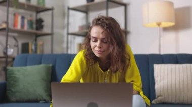 Smiling young woman in casual attire sitting on comfortable sofa and using laptop for online study or work. Concept of people, gadgets and domestic lifestyles.