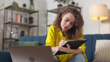 Oturma odasındaki kahve masasında dizüstü bilgisayarda çalışan güzel genç bir kadın. Çekici beyaz kadın, rahat bir kanepede otururken günlük giysiler içinde panoya yazı yazıyor..