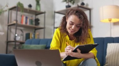 Modern dizüstü bilgisayarın önünde elinde dosyayla oturan beyaz bir kız öğrenci. Kıvırcık saçlı pozitif genç bir kadın online çalışırken not alıyor..