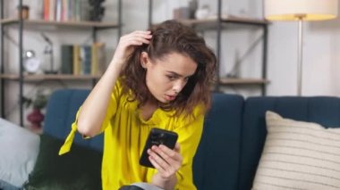 Pretty young woman with beautiful make up looking at her reflection on smartphone and fixing her curly brown hair. Attractive female sitting on comfortable couch and using mobile.