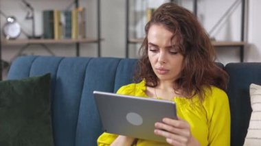 Kafkasyalı ciddi bir kadın rahat bir odada kanepede oturup dijital tablet üzerinde çalışıyor. Genç bir kadın, e-postaları kontrol ediyor ya da modern bir cihazda mesaj yazıyor..