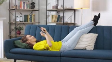 Beautiful relaxed woman leaning on comfortable sofa cushion, using digital smartphone and enjoying day off. Leisure time at home with modern gadgets.