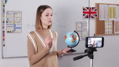 Female english tutor using modern smartphone for recording video lesson. Woman turning globus and translating new words for students during distance learning.
