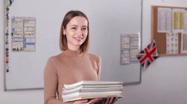 Smiling caucasian tutor holding stack of grammar books before English lesson in modern classroom. Positive charming woman in casual wear preparing for next lecture.