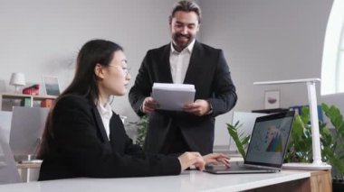 Confident young woman getting paper with good news from executive while working on laptop at office. Bearded male boss hugging and congratulating female employee with achievement.