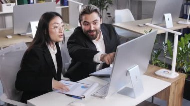 Happy multiracial business analysts sitting at desk with computer in modern office. Caucasian man and asian woman looking at monitor with financial graphics.