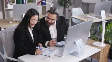 Caucasian businessman and asian businesswoman discussing sales report on clipboard at office desk. Two company coworkers planning strategy for good profit in front of computer.