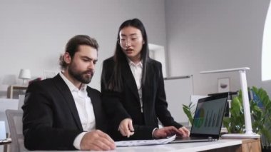 Angry asian female boss throwing documents of financial report to employee and pointing mistakes. Bearded man sitting at desk with laptop in bright office trying to find excuses.