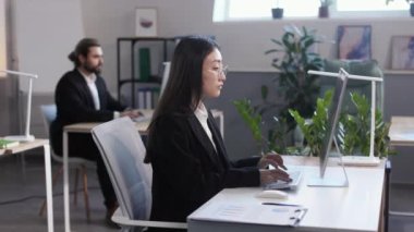Side view of asian young woman in formal wear typing on keyboard and looking on monitor with focused facial expression. Blur background of caucasian male coworker at office.