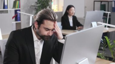 Tired caucasian man wearing black suit suffering from headache while sitting at workplace with modern pc. Concept of overworked, exhaustion and stress.