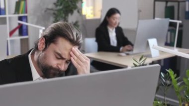 Frustrated businessman massaging forehead while suffering from strong migraine at office. Caucasian man sitting at desk and feeling tired from long work on modern pc.