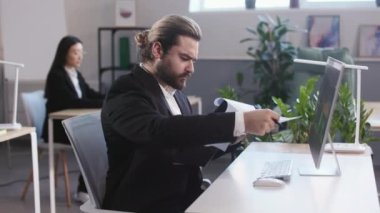 Bearded male trading specialist sitting at desk with modern computer and reading documents on clipboard. Young man in formal suit checking execution of sales plan.