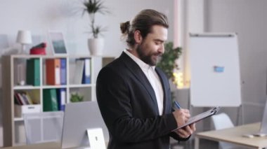 Portrait of bearded man in formal suit holding clipboard and looking at camera with smile. Male worker with long hair making notes before business meeting at office.
