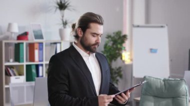 Friendly staff member in formal suit looking at camera with smile while preparing for business meeting. Young man holding clipboard with important speech at bright office room.