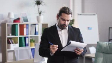 Satisfied young marketer in suit posing on camera while holding clipboard with document at bright office. Bearded male making notes in business plan and feeling pleasure about new success strategy.