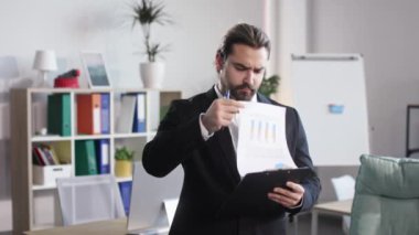 Portrait of pensive employer writing notes on clipboard while standing in modern meeting room. Caucasian man in formal wear searching mistake in documents before meeting with partners.