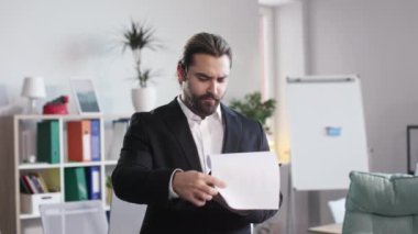 Young office worker standing in middle of agency meeting room and holding clipboard with important documents. Bearded man shaking head with approval while reading strategy plan.